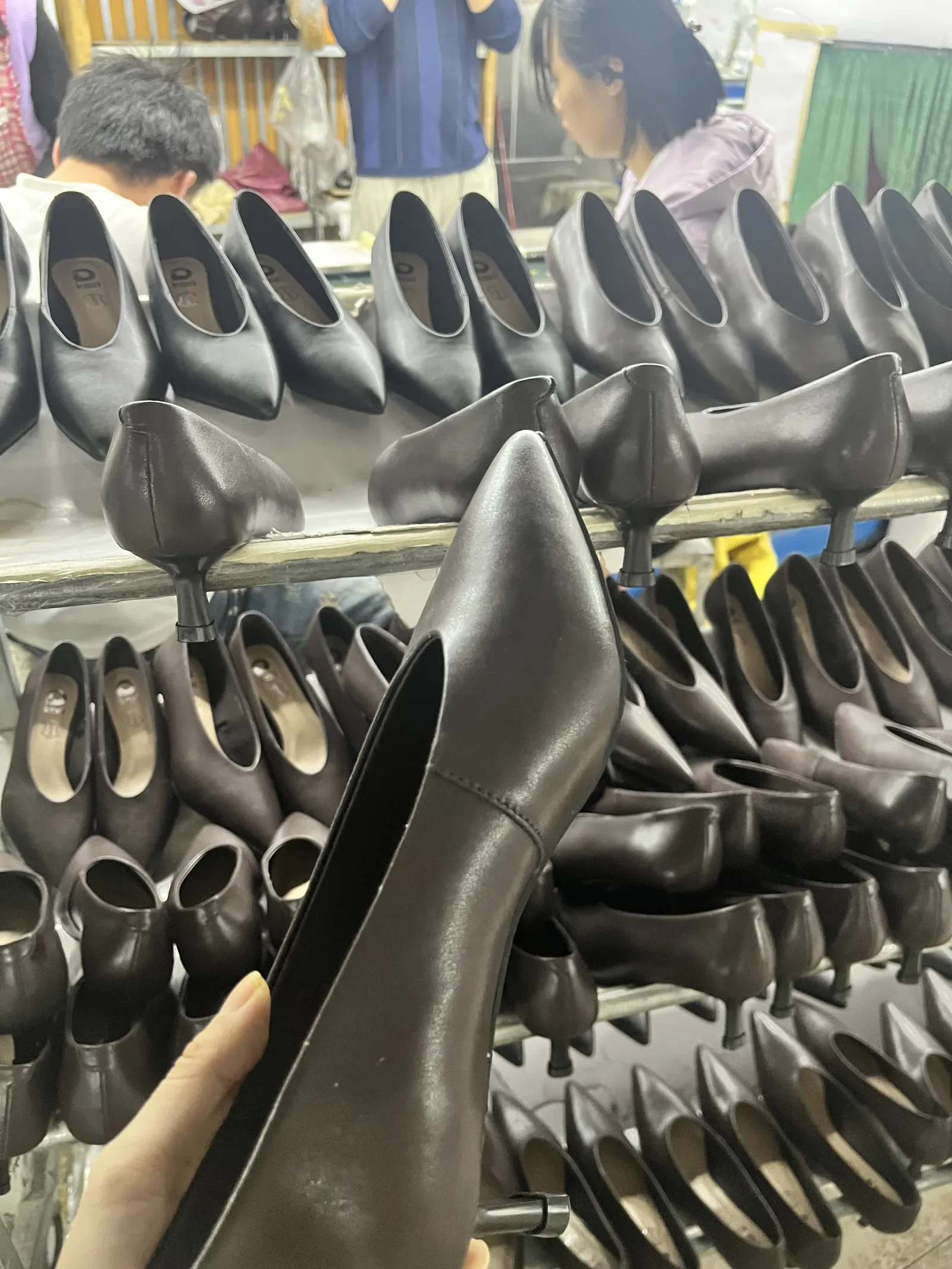 Hand inspecting a brown pointed-toe dress shoe against racks of finished pairs during sample approval